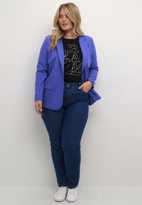 Lila blézer sima anyagból, fekete grafikás pólóval és kék skinny farmerral párosítva. Fehér sportcipő egészíti ki az öltözetet.