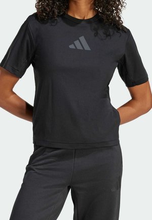 Femme aux cheveux bouclés portant un t-shirt noir à manches courtes et un pantalon noir assorti avec un logo discret sur les deux.