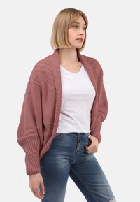 Cardigan tricoté en rose poussiéreux avec un motif texturé, des manches larges et des poignets côtelés, associé à un t-shirt blanc et un jean bleu.