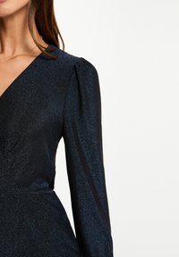 Une femme portant une robe portefeuille longue à manches longues d'un bleu foncé, scintillante, avec des épaules légèrement bouffantes et un délicat collier en chaîne dorée.