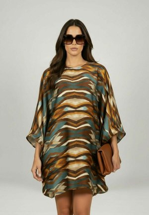 Vrouw draagt oversized bruine, turquoise en beige jurk met patroon, grote donkere zonnebril, houdt een bruine clutch vast tegen een effen achtergrond.