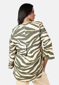 Femme aux longs cheveux noirs portant un haut oversize rayé zèbre vert et beige avec des manches trois-quarts, de profil avec les mains dans les poches de son pantalon beige.