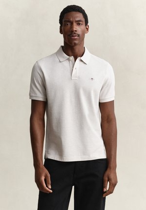 Man met kort krullend haar en snor, gekleed in een lichtbeige poloshirt en zwarte broek, staand tegen een effen achtergrond.