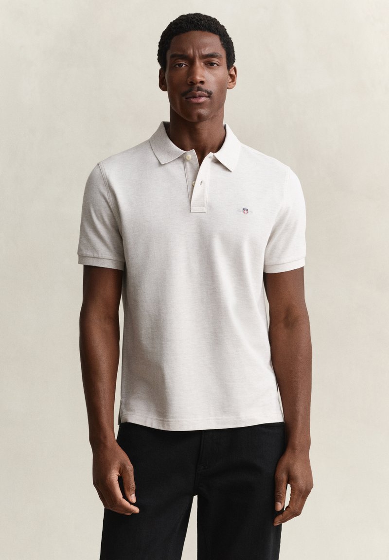 Homme aux cheveux courts et bouclés et à la moustache, portant un polo beige clair et un pantalon noir, debout devant un fond uni.