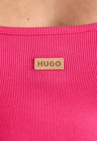 Ribbota rozā blūze ar kvadrātveida dekoltē un brūnu ādas etiķeti ar iepresētu "HUGO", kas šūta pie priekšpuses.