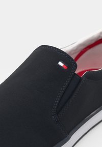 Námořnické modré slip-on tenisky s bílým, červeným a námořnickým logem na svršku, bílou podrážkou s černým pruhem a červeným vnitřním podšívkou.