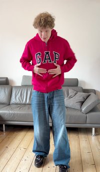 Camisola com capuz vermelha e fecho de correr, com o logótipo "GAP" em azul-marinho, usada com jeans de perna larga azul-claro. Sapatos pretos, em pé contra uma parede de cor clara.