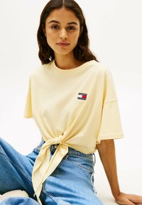 T-shirt di cotone giallo con dettaglio annodato in vita e una piccola patch logo multicolore sul lato sinistro, abbinato a jeans blu a vita alta.