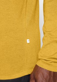 Senapsgul långärmad stickad tröja med en strukturerad finish. Har en liten vit märkeslapp på den vänstra sömmen.