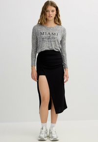 Camiseta gris de manga larga con la inscripción "Miami", combinada con una falda negra fruncida que tiene una abertura alta en el muslo. La modelo lleva zapatillas deportivas grises.