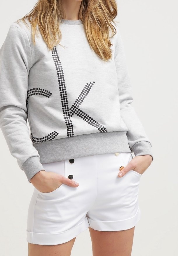 Sudadera gris con el logo "CK" en tachuelas negras y ribete gris; combinada con pantalones cortos blancos que presentan botones dorados y negros, con bolsillos visibles.