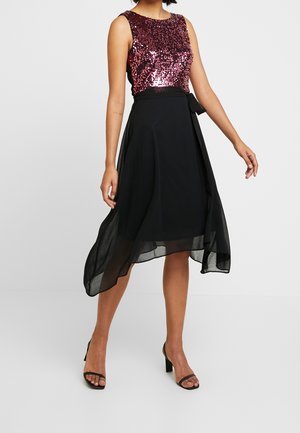 Kobieta ubrana w sukienkę bez rękawów z ciemnoróżową cekinową górą i asymetryczną czarną spódnicą z szyfonu, w zestawie z czarnymi sandałami na obcasie.