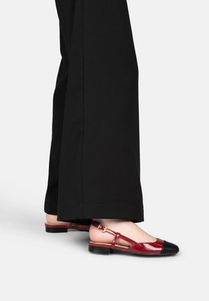 Personne portant un pantalon noir à jambes larges et des ballerines rouges slingback avec bouts noirs, debout sur une surface blanche.