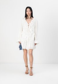 Λευκό φόρεμα με μακριά μανίκια, φουσκωτή μπούστα και λεπτομέρεια με κόμπο στο λαιμό. Το μοντέλο κρατάει μία μπλε τσάντα, φοράει σαγιονάρες σε μπεζ απόχρωση.