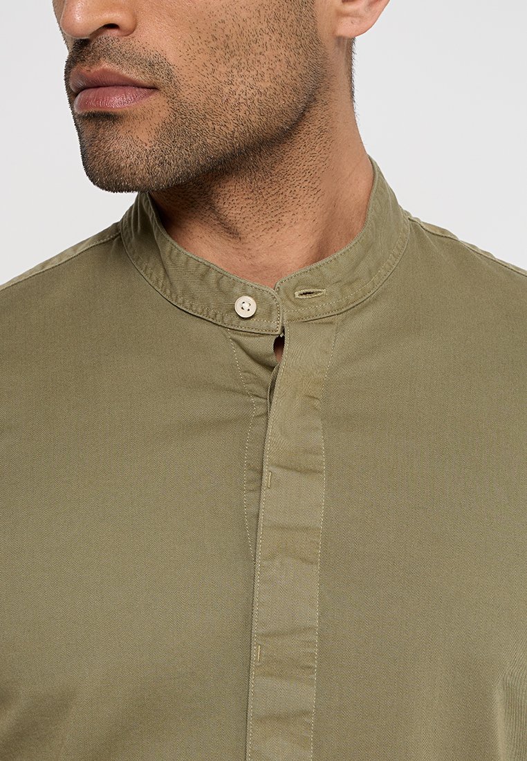 Eine olivgrüne Bluse mit Stehkragen, die eine zentrale Knopfleiste hat, die mit einem kleinen Knopf verschlossen wird, aus Baumwollmaterial mit glatter Textur.