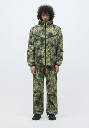 Giacca e pantaloni in un motivo mimetico nei toni del verde. Caratteristiche includono un colletto alto, tasche con zip e polsini elasticizzati.