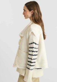 Crème-kleurige jas van zachte stof, met een sjawl-kraag, twee voorzakken en zwarte gestreepte accenten op de mouwen.