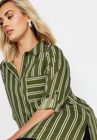 Camicia verde con righe verticali bianche, chiusura a bottoni sul davanti, colletto e una tasca sul petto. Maniche lunghe arrotolate ai polsi, tessuto testurizzato.