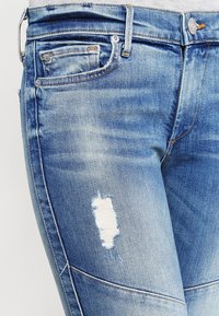 Jeans de denim azul claro con un corte regular, desgastes visibles y un área desgastada en el muslo izquierdo. Incluye detalles de hardware en color cobre.