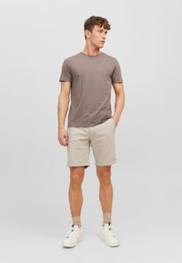 Camiseta de manga corta marrón combinada con pantalones cortos beige. El modelo lleva zapatillas blancas con detalles azules y calcetines beige. Diseño sencillo y casual.