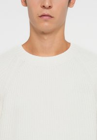 Balts adījuma džemperis ar ribotu tekstūru, apaļu kakla izgriezumu un raglanu piedurknēm, piedāvājot pieguļošu siluetu.