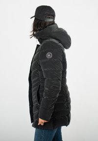 Manteau en duvet noir avec capuche, présentant un motif matelassé horizontal, des poches latérales et un logo sur la manche gauche. Stylisé avec un jean bleu.