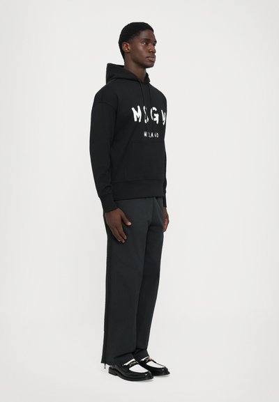 Modello maschile in piedi, vestito con una felpa nera con il testo "MSGM Milano", pantaloni larghi scuri, calze bianche e mocassini bianco e nero con catena dorata.