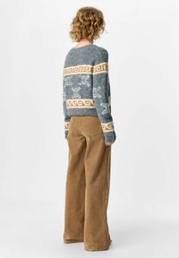 Person mit lockigem Haar, der einen grauen, gemusterten Pullover und weite, braune Cordhosen trägt, steht mit dem Rücken zu einer weißen Wand.