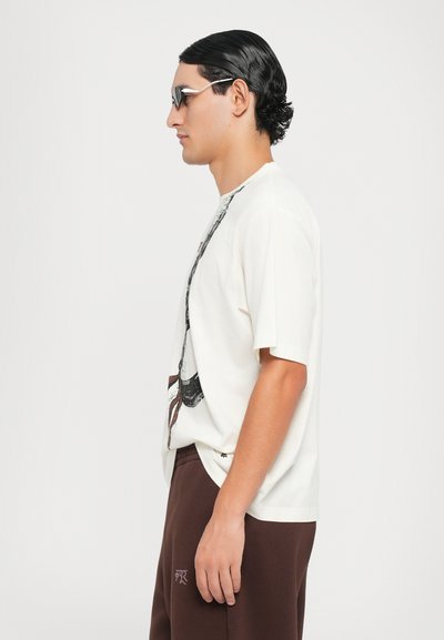 Beige Grafik-T-Shirt mit kurzen Ärmeln, versehen mit einem schwarzen und roten Druckdesign, kombiniert mit braunen lässigen Hosen. Akzentuiert mit stylischen Sonnenbrillen.
