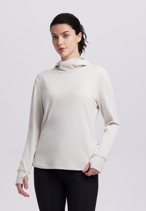 Kvinde med mørkt hår iført en cremefarvet langærmet hættetrøje med tommelfingerhuller og sorte bukser, stående foran en lysegrå baggrund.