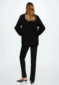 Blazer noir surdimensionné à manches longues, associé à un pantalon noir ajusté et des escarpins noirs à bout pointu avec un motif décoratif.