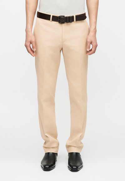 Άνδρας που φοράει μπεζ παντελόνι φόρμας με μαύρη ζώνη και μαύρα δερμάτινα παπούτσια, στέκεται μπροστά σε απλό λευκό φόντο.