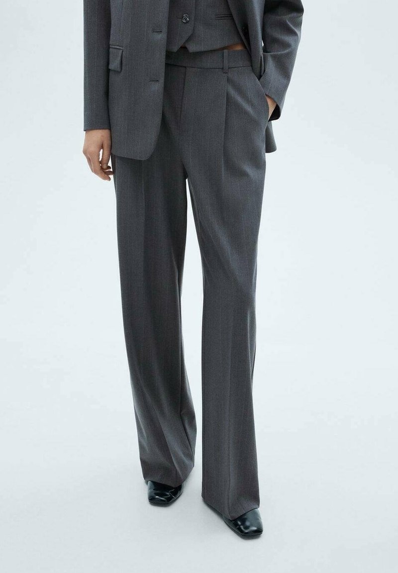 Personne portant un pantalon gris à fines rayures et un blazer assorti, avec une main dans la poche et des chaussures noires à bout pointu visibles.