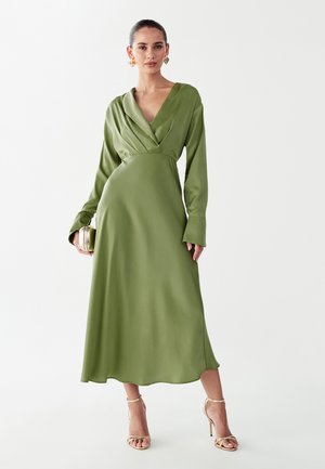 Saténové zelené šaty s hlbokým výstrihom, dlhými rukávmi a priliehavým pásom, so splývavou sukňou a jemným leskom. Zahŕňa zlaté doplnky.