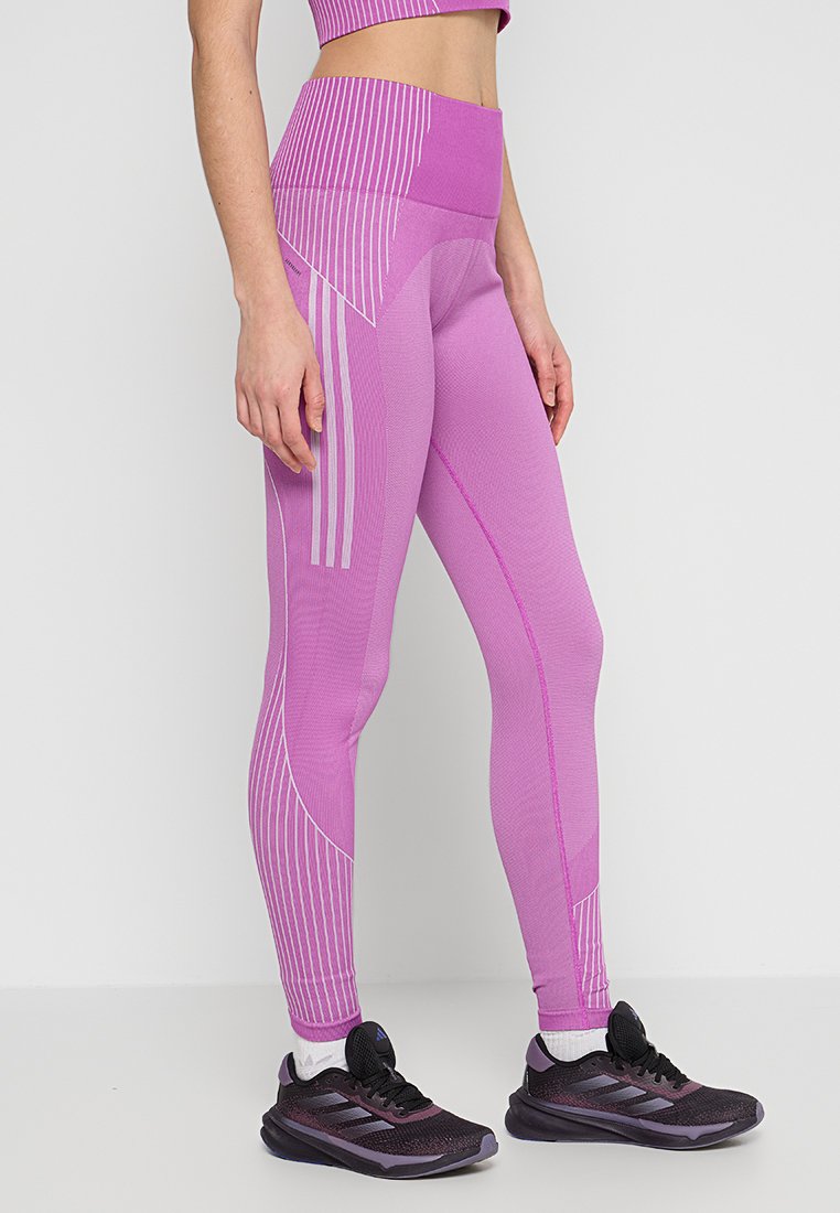 Leggings violets à taille haute en tissu texturé, avec des rayures blanches verticales et une silhouette ajustée, associés à des chaussures de sport noires.
