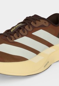 Καφέ παπούτσι για τρέξιμο Adidas με λευκές ρίγες, σκούρα καφέ κορδόνια και μπεζ σόλα με την ένδειξη "Lightstrike Pro."