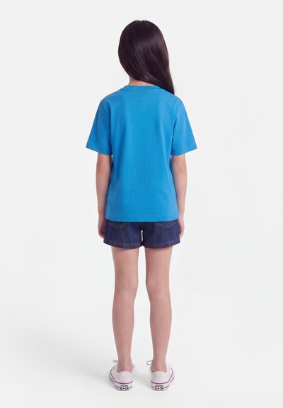 Dijete s dugom tamnom kosom, odjeveno u plavu majicu, tamne traper kratke hlače i bijele tenisice, stoji okrenuto leđima na bijeloj pozadini.