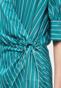 Τυρκουάζ φόρεμα με λευκές κάθετες ρίγες, δεμένο στη μέση με κόμπο, που αποκαλύπτει μέρος από ένα κοντό μανίκι με μανσέτα και το χέρι.