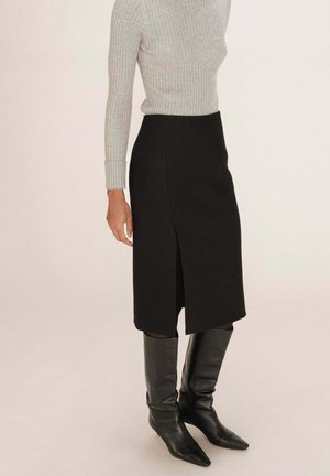 Jupe midi noire avec fente sur le côté, fabriquée en tissu texturé. Associée à un pull en laine côtelé gris clair et des bottes en cuir noires montantes.