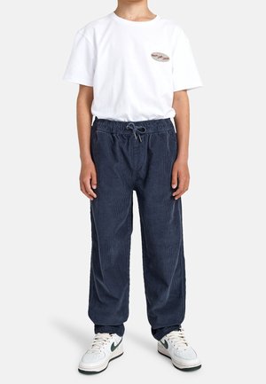 Enfant portant un t-shirt blanc avec un petit logo à l'avant, un pantalon en velours côtelé bleu foncé avec cordon de serrage, et des baskets blanches avec un logo swoosh noir.