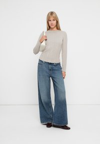 Lichtgrijs geribbeld langemouwen shirt gecombineerd met een wijde blauwe denim jeans. Het model houdt een kleine witte tas vast en draagt glanzende donkere schoenen.
