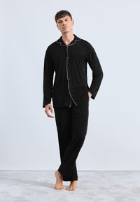 Conjunto de pijama negro hecho de tejido suave, con una camisa de botones con ribetes blancos y pantalones a juego, mostrado en un modelo descalzo.