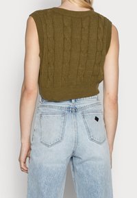 Woman wearing olive green sleeveless cable knit sweater vest and light blue high-waisted denim jeans, shown from the back.