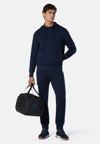 Sudadera de marina y pantalones jogger de tejido suave, con un logo en el pecho. Hombre sostiene un bolso de deporte negro con logo, vistiendo zapatillas oscuras.