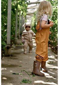 Kind draagt een okergele waterdichte overal en bruine rubberen laarzen. Een ander kind in een beige outfit met konijnenoren staat achter hen. Groene bladeren omringen hen.