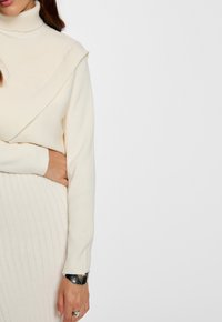 Maglione a coste di colore crema con colletto alto e design frontale a strati, abbinato a una gonna a coste coordinata. Presenta un bracciale argentato al polso.