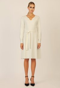 Cremefarbenes, langärmeliges Kleid mit V-Ausschnitt und einem gebundenen Taillengürtel, A-Linien-Silhouette, glatter Textur, knielanger Saum, kombiniert mit schwarzen Absätzen.