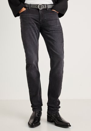 Jeans de mezclilla negra con corte slim, que presentan una textura sutil y una cintura de altura media. Accesorios con un cinturón negro con tachuelas y botas negras elegantes.