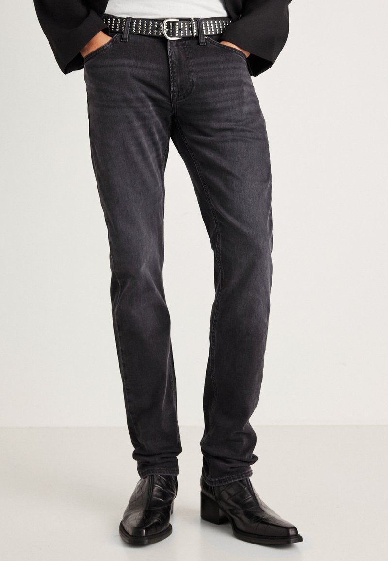 Mustad denim püksid slim-fit lõikega, millel on õrn tekstuur ja keskmise kõrgusega vöökoht. Aksessuaariks on studsiga must vöö ja stiilsed mustad saapad.