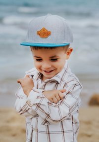 Szürke sapka kék karimával és narancssárga logóval, fehér kockás inggel, amely barna és kék vonalakkal díszített. A gyermek játékosan gesztikulál.